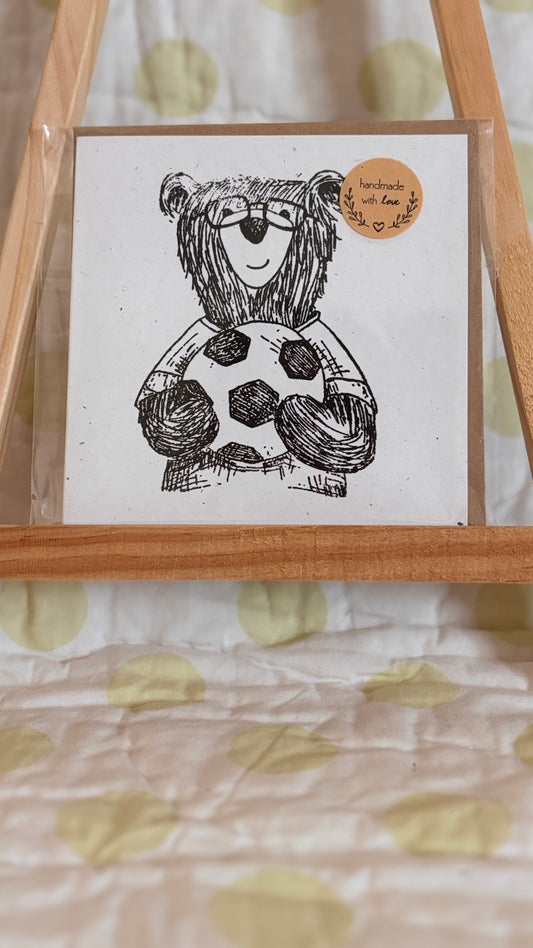 Card with a bear illustration holding a soccer ball, framed by a wooden stand on a patterned background.